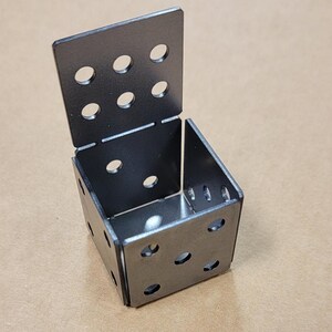 May include: A metal cube made of four square pieces with holes drilled in each piece. The cube is open on one side.