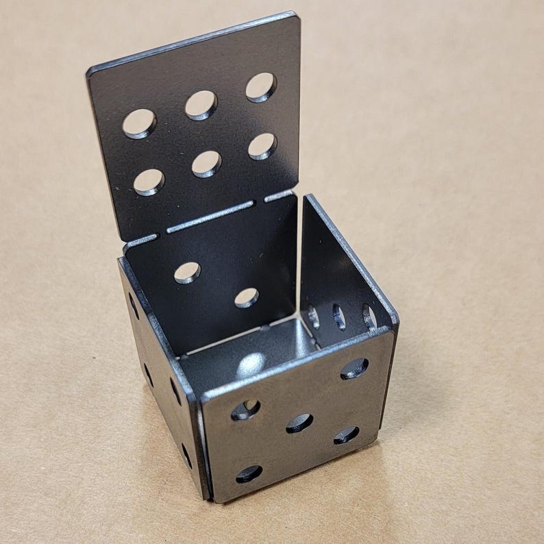 May include: A metal cube made of four square pieces with holes drilled in each piece. The cube is open on one side.