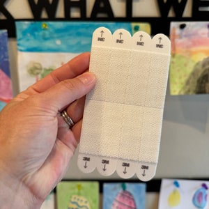 May include: A white, rectangular piece of 3M Command Strip with four strips of adhesive. The strips are labeled "3M" and have arrows pointing down. The top of the strip has four strips labeled "WC" with arrows pointing up.