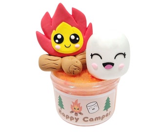May include: A clear plastic container filled with orange slime, topped with a clay figurine of a campfire with a smiling face and a white marshmallow with a smiling face. The container has a label that says "Happy Camper" with a small campfire and marshmallow graphic.
