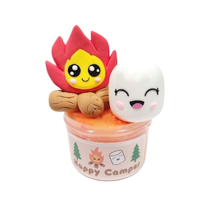 May include: A clear plastic container filled with orange slime, topped with a clay figurine of a campfire with a smiling face and a white marshmallow with a smiling face. The container has a label that says "Happy Camper" with a small campfire and marshmallow graphic.