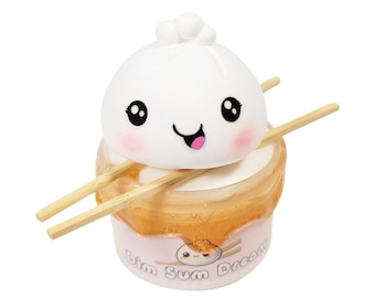 May include: A white, cartoon-style dim sum dumpling with a smiling face sits on top of a clear container with a pink and gold design. The container has the text "Dim Sum Dream" on the front. Two wooden chopsticks are placed on top of the dumpling and container.