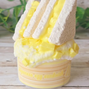 May include: A miniature Japanese egg sandwich, featuring fluffy white bread and a creamy yellow filling. The sandwich sits atop a yellow and white container with the words "Japanese egg sandwich" printed on it. The food item is a dessert or novelty item.