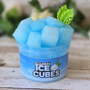 May include: A clear plastic container filled with blue, cube-shaped slime, topped with a green leaf decoration. The container has a blue label with the text "Ice Slimers Ice Cubes". The background is a blurred wooden surface.