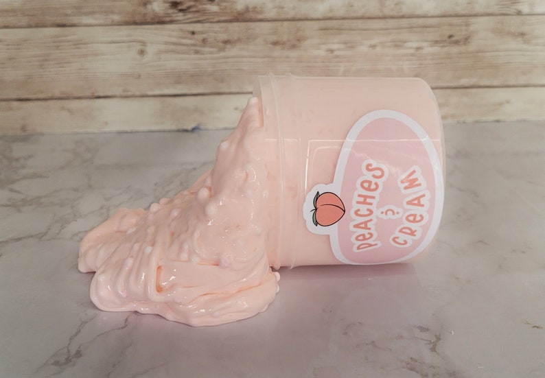 Puede incluir: Un recipiente de slime color melocot&oacute;n con acentos blancos texturizados. El recipiente tiene una etiqueta que dice "Peaches & Cream" con un gr&aacute;fico de melocot&oacute;n. El slime est&aacute; parcialmente derramado del recipiente.