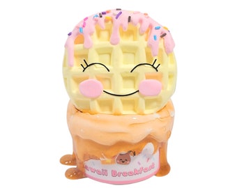 May include: A yellow waffle with pink icing and sprinkles, a smiling face, and pink cheeks. The waffle is sitting on top of a brown, melted syrup-like substance. The base of the syrup has a pink label that says "Kawaii Breakfast".