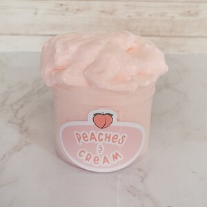 Puede incluir: Un recipiente de pl&aacute;stico transparente lleno de slime rosa claro. El recipiente tiene una etiqueta con un gr&aacute;fico de melocot&oacute;n y las palabras "Peaches & Cream" en un dise&ntilde;o rosa y blanco. El slime es esponjoso y texturizado.
