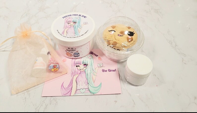 May include: A pink and green slime jar with a cartoon girl on the label. The jar is labeled "Mochi Fun Slime". The slime is packaged with a cookie, a small jar of slime, and a pink card with a cartoon girl on it. The card says "All the time!"