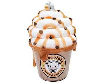 May include: A chocolate cow-shaped cup with a white whipped cream topping, drizzled with caramel and chocolate chips. The cup has a brown and yellow logo that says "Chocolate Cowpuccino".