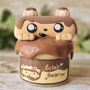May include: A whimsical jar with a bear-shaped eclair design. The bear has large black eyes and small ears. The jar is brown and tan with the words "Éclair Bear" on the front.