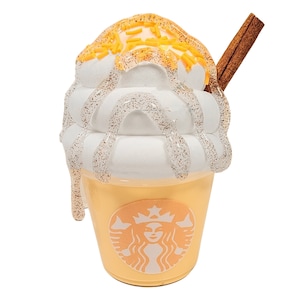 Könnte beinhalten: Eine dekorative Kaffeetasse mit weißem Schlagsahne-Topping, beträufelt mit glänzendem Sirup. Die Tasse ist hellorange mit dem Starbucks-Logo. Ein Zimtstange ist in der Schlagsahne platziert.
