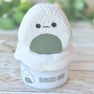 May include: A whimsical, white, textured onigiri-shaped item with black eyes and a smiling mouth, atop a white cloud-like base. The item sits on a small container with the text "Onigiri Cutie" and a graphic of an onigiri.