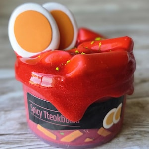 May include: A small, pink container with a red, glossy topping, resembling a Korean dish. The container is labeled "Spicy Tteokbokki" and is decorated with egg and rice cake illustrations. Two egg slices sit atop the red topping.