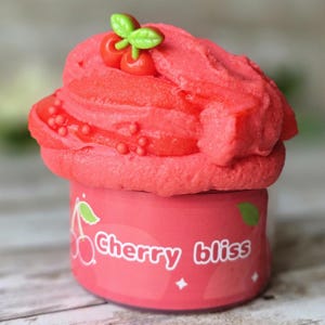 May include: A vibrant pink container of "Cherry bliss" slime, topped with a swirl of textured pink slime and cherry-shaped embellishments. The container is adorned with a cherry illustration and the words "Cherry bliss".