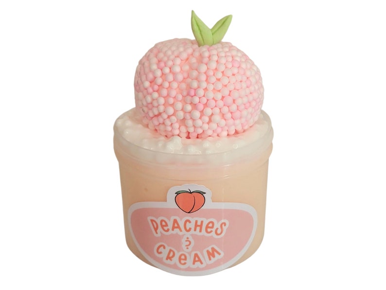 Puede incluir: Un recipiente de pl&aacute;stico transparente lleno de slime color melocot&oacute;n, coronado con una esfera rosa hecha de peque&ntilde;as bolas y un acento de hoja verde. La etiqueta dice "Peaches & Cream" con un gr&aacute;fico de melocot&oacute;n.