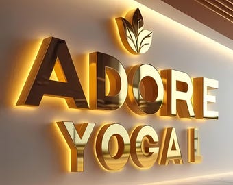 May include: A gold-colored sign with the words "ADORE YOGA" in raised letters with a leaf design above the words.