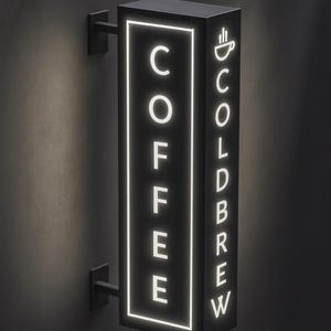 May include: A black, rectangular coffee shop sign with the word "COFFEE" vertically stacked on one side and "COLD BREW" on the other. The words and a coffee cup icon are illuminated with a bright white light. The sign is mounted on a dark wall.