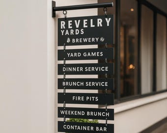 May include: Black rectangular sign with white text hanging from a black metal rod. The sign reads "REVELRY YARDS BREWERY" and lists services including "YARD GAMES," "DINNER SERVICE," and "BRUNCH SERVICE." Additional text includes "FIRE PITS," "WEEKEND BRUNCH," and "CONTAINER BAR."