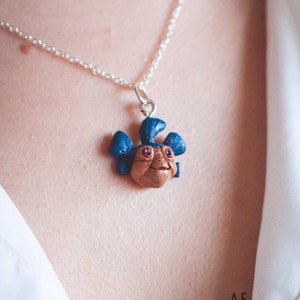 Könnte beinhalten: Halskette mit einem Anhänger, der ein braunes und blaues Wesen mit roten Augen zeigt, an einer feinen Silberkette. Der Anhänger hat ein lächelndes Gesicht und blaue, ohrenartige Anhängsel. Handgefertigtes Sammlerstück.