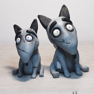 May include: Two gray, stitched, dog figurines with black ears and eyebrows. The dogs have large, white eyes and are sitting on a light gray surface.