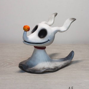 May include: A white ceramic figurine of Zero, the ghost dog from The Nightmare Before Christmas, with a small orange pumpkin on his nose. Zero has black eyes and a red collar. The figurine is sitting on a light wood surface.