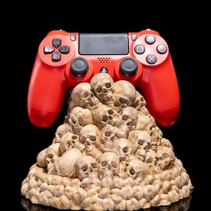 May include: A red video game controller sits atop a skull-shaped stand. The stand is made of a light brown material and is covered in small, detailed skulls.