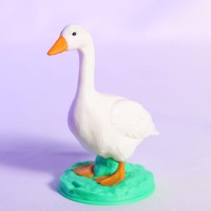May include: A white goose figurine with an orange beak and feet, standing on a small, green base. The goose has a long neck and is facing to the left. The background is a gradient of purple and white.