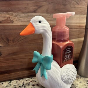 May include: A white goose-shaped soap dispenser holder with an orange beak and a teal bow. The holder is designed to hold a brown bottle of hand soap. The soap bottle has a pink pump.