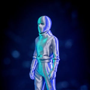 May include: A silver 3D printed figurine of a person wearing a full-face mask and a hooded jumpsuit. The figurine is standing with its arms at its sides and is illuminated by blue and green lights.