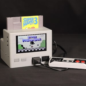 Puede incluir: Una consola de videojuegos retro gris con una pantalla integrada que muestra la pantalla de título de Super Mario Bros. Un cartucho de juego Super Mario Bros. 3 está insertado en la ranura superior. Un mando rectangular blanco y negro con dos botones rojos está conectado a la consola mediante un cable negro.