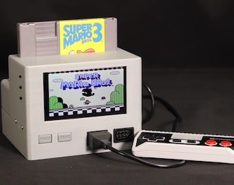 May include: A gray retro-style video game console with a built-in screen displaying the Super Mario Bros. title screen. A Super Mario Bros. 3 game cartridge is inserted into the top slot. A rectangular white and black controller with two red buttons is connected to the console via a black cable.