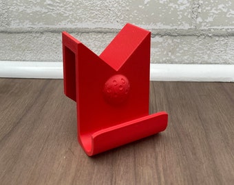 May include: A red plastic phone holder with a golf ball design. The holder is designed to be mounted on a wall.