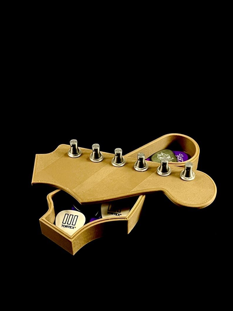 Puede incluir: Una caja de madera con forma de clavijero de guitarra con clavijas plateadas. La caja est&aacute; abierta, revelando p&uacute;as de guitarra con la marca "Tortex". La caja es de madera marr&oacute;n claro sobre un fondo negro.