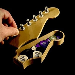 Puede incluir: Un soporte de p&uacute;as de guitarra de madera con forma de clavijero, abierto para revelar una variedad de p&uacute;as de guitarra. El soporte es de color marr&oacute;n claro, con clavijas plateadas. Las p&uacute;as de guitarra son de varios colores, incluyendo blanco, morado y negro, con la marca "Tortex" visible.