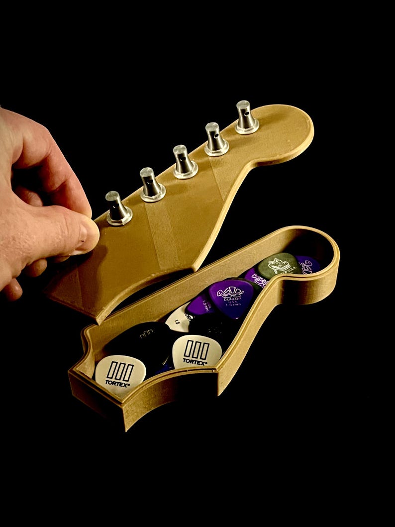 Puede incluir: Un soporte de p&uacute;as de guitarra de madera con forma de clavijero, abierto para revelar una variedad de p&uacute;as de guitarra. El soporte es de color marr&oacute;n claro, con clavijas plateadas. Las p&uacute;as de guitarra son de varios colores, incluyendo blanco, morado y negro, con la marca "Tortex" visible.