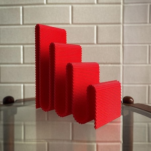 May include: Four bright red, textured, folded rectangles of varying heights are arranged in a descending stair-step pattern. The objects are on a reflective surface, with a light-coloured brick wall in the background.