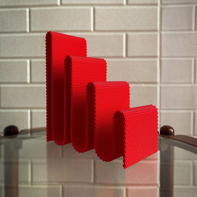 May include: Four bright red, textured, folded rectangles of varying heights are arranged in a descending stair-step pattern. The objects are on a reflective surface, with a light-coloured brick wall in the background.