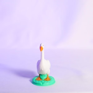 May include: A white goose figurine with an orange beak and feet stands on a green base. The goose is centered against a soft purple background, creating a whimsical and charming aesthetic. The figurine is a decorative item.