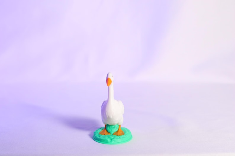 May include: A white goose figurine with an orange beak and feet stands on a green base. The goose is centered against a soft purple background, creating a whimsical and charming aesthetic. The figurine is a decorative item.