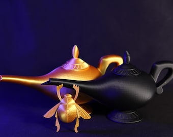Könnte beinhalten: Eine goldfarbene und eine schwarze Dschinn-Lampe werden auf einer dunkelblauen Oberfläche präsentiert. Vor den Lampen befindet sich eine goldfarbene Skarabäus-Figur. Die Lampen haben gebogene Ausgüsse und Griffe.