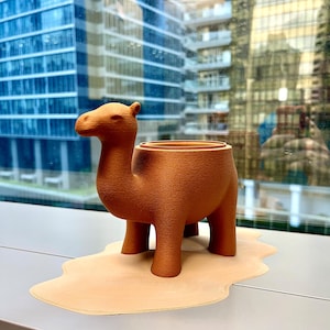 May include: A terracotta-colored camel-shaped planter with a textured surface. The planter has a large opening for plants and sits on a beige, desert-like mat. The background features a cityscape.