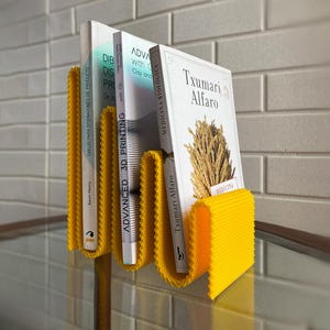 May include: A vibrant yellow book holder in a wavy design, holding three books. The books feature white covers with text in Spanish and English. The holder is made of a textured material and is on a glass surface.