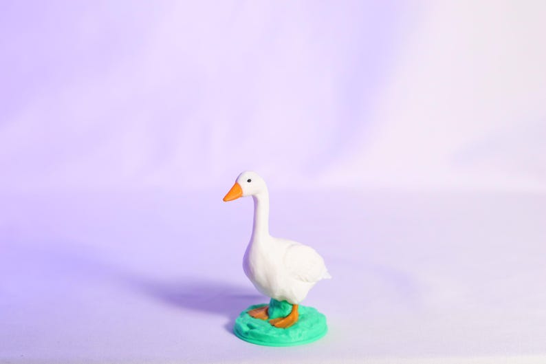 May include: A white goose figurine with an orange beak and feet, standing on a small, green base. The goose has a long neck and is facing to the left. The background is a gradient of purple and white.