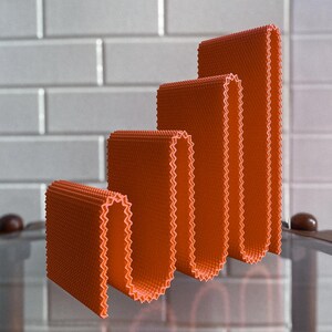 May include: Four orange, folded, rectangular objects of varying heights, arranged in a stepped pattern. Each object has a textured surface and a zig-zag edge. The background features a white tiled wall.