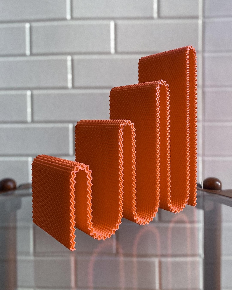 May include: Four orange, folded, rectangular objects of varying heights, arranged in a stepped pattern. Each object has a textured surface and a zig-zag edge. The background features a white tiled wall.