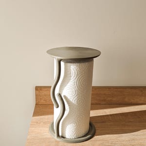 Puede incluir: Un moderno portarrollos de papel de cocina con base y parte superior de hormigón gris, y un diseño curvo. El soporte está diseñado para sostener un rollo de toallas de papel blancas con un patrón circular. El soporte está sobre una superficie de madera.