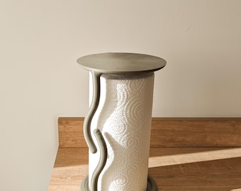 May include: A modern paper towel holder with a gray concrete base and top, and a curved design. The holder is designed to hold a roll of white paper towels with a circular pattern. The holder is on a wooden surface.