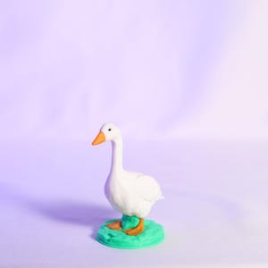May include: A white goose figurine with an orange beak and feet stands on a green base. The goose has a long neck and is set against a soft purple background. The figurine is a decorative item.