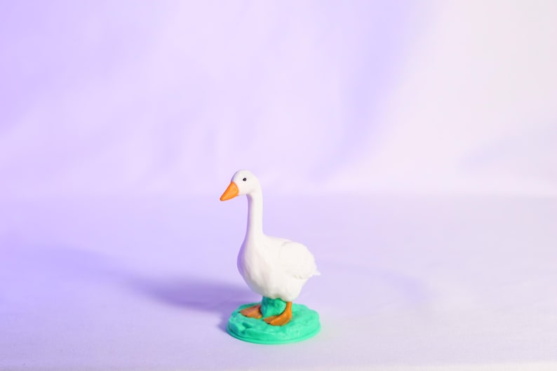 May include: A white goose figurine with an orange beak and feet stands on a green base. The goose has a long neck and is set against a soft purple background. The figurine is a decorative item.