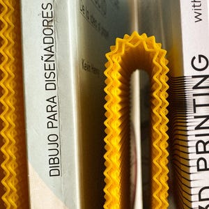 May include: A yellow, wavy-edged 3D-printed bookend supporting several books. The books have white covers with text in Spanish and English. The bookend's bright colour contrasts with the book covers, creating a visually appealing arrangement.